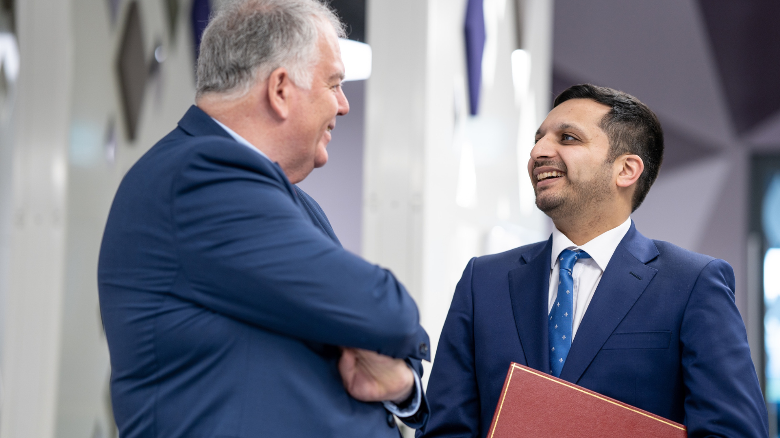 Two men in blue suits are engaged in a cheerful conversation indoors.
