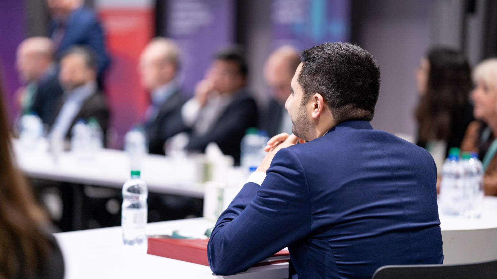 A man in a blue suit sits at a conference table, attentively listening to a speaker.