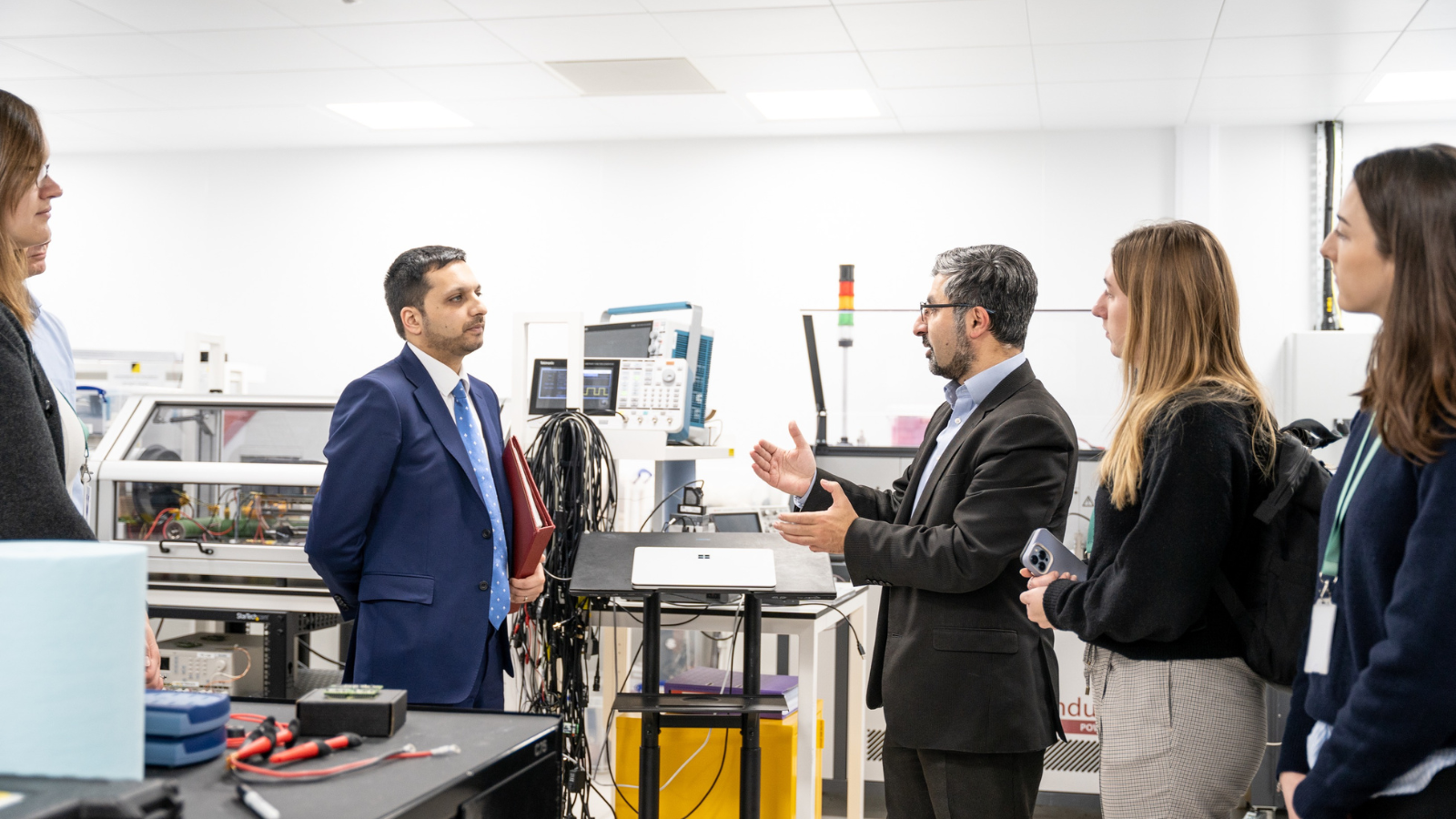A group of people, some in formal attire, are having a discussion in a high-tech laboratory.