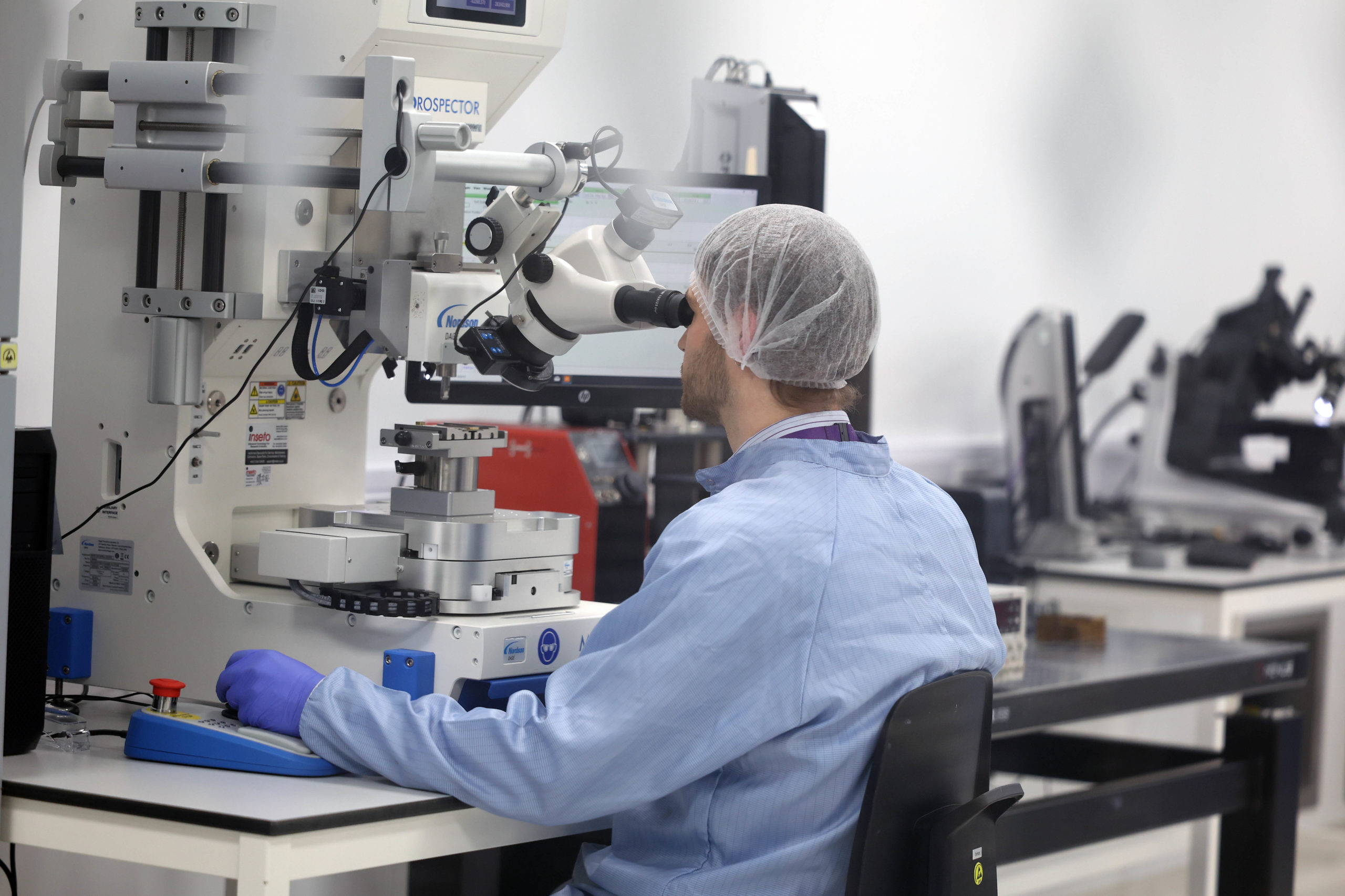 A scientist in a lab coat and hair net uses a microscope at a laboratory workbench. - CSA Catapult