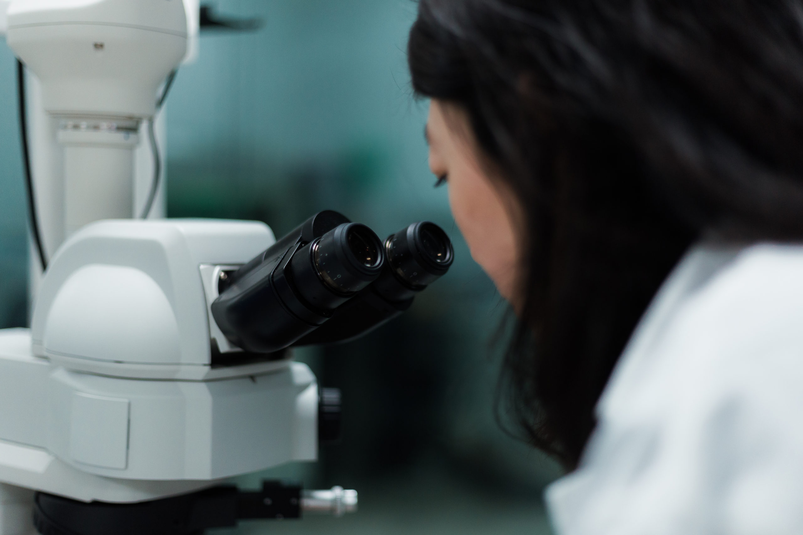 Person in a lab coat looking through the eyepieces of a white laboratory microscope. - CSA Catapult
