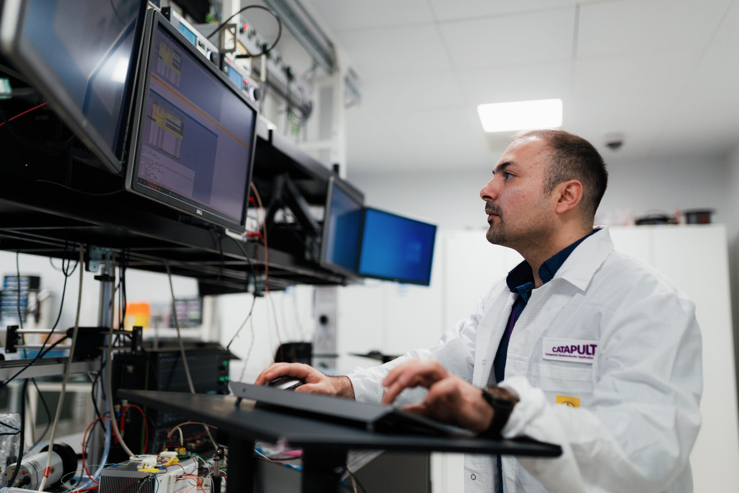 A man in a lab coat works on a computer in a laboratory with multiple monitors. - CSA Catapult