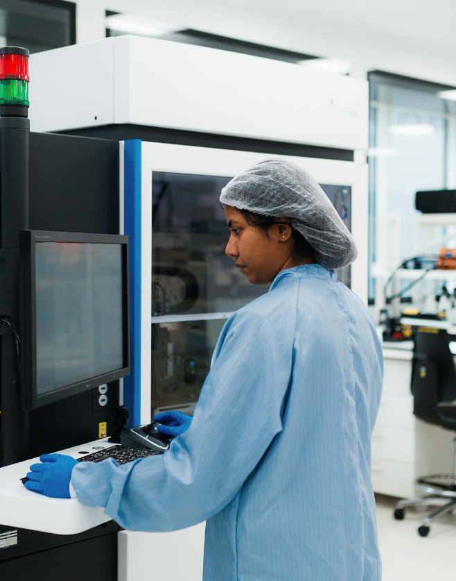 A person wearing a blue lab coat, hair net, and gloves operates a machine in a bright, modern lab with glass walls and various scientific equipment. The lab is well-lit with natural and artificial lighting. - CSA Catapult