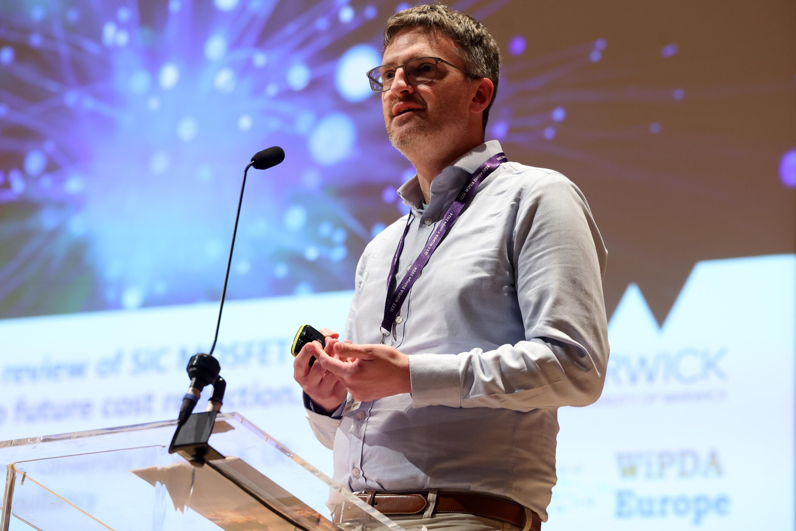 A man stands at a podium speaking into a microphone during a conference, with a presentation screen behind him. - CSA Catapult