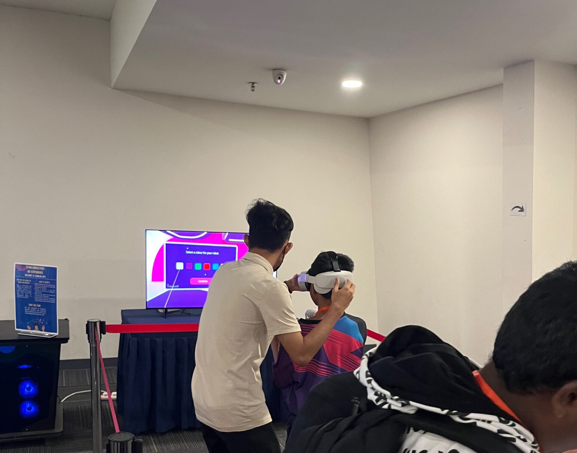 A person helps another try a VR headset in a room where a TV shows a T-shaped digital interface. - CSA Catapult