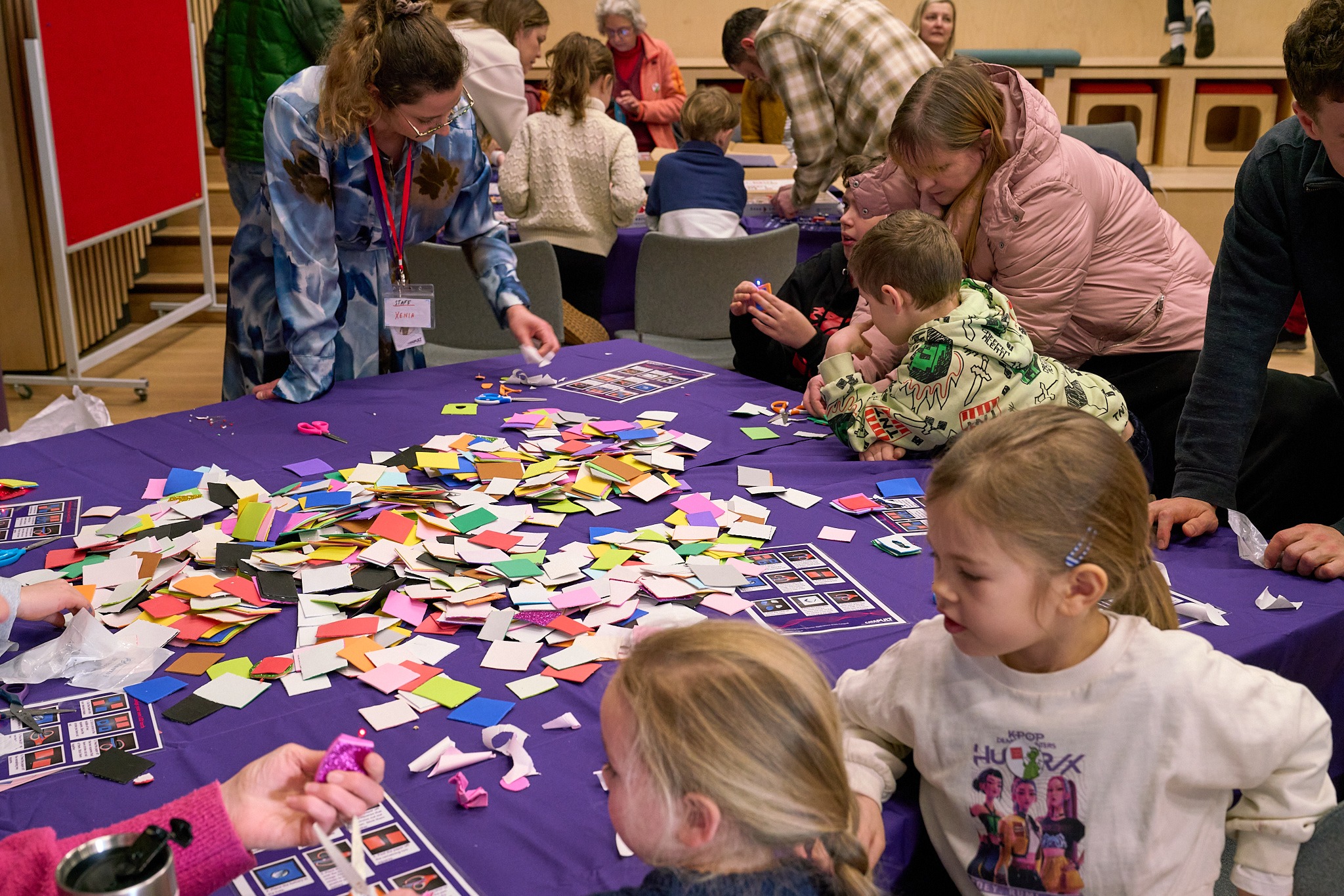Children and adults gather around a table covered with colorful paper squares, engaging in a craft activity. - CSA Catapult