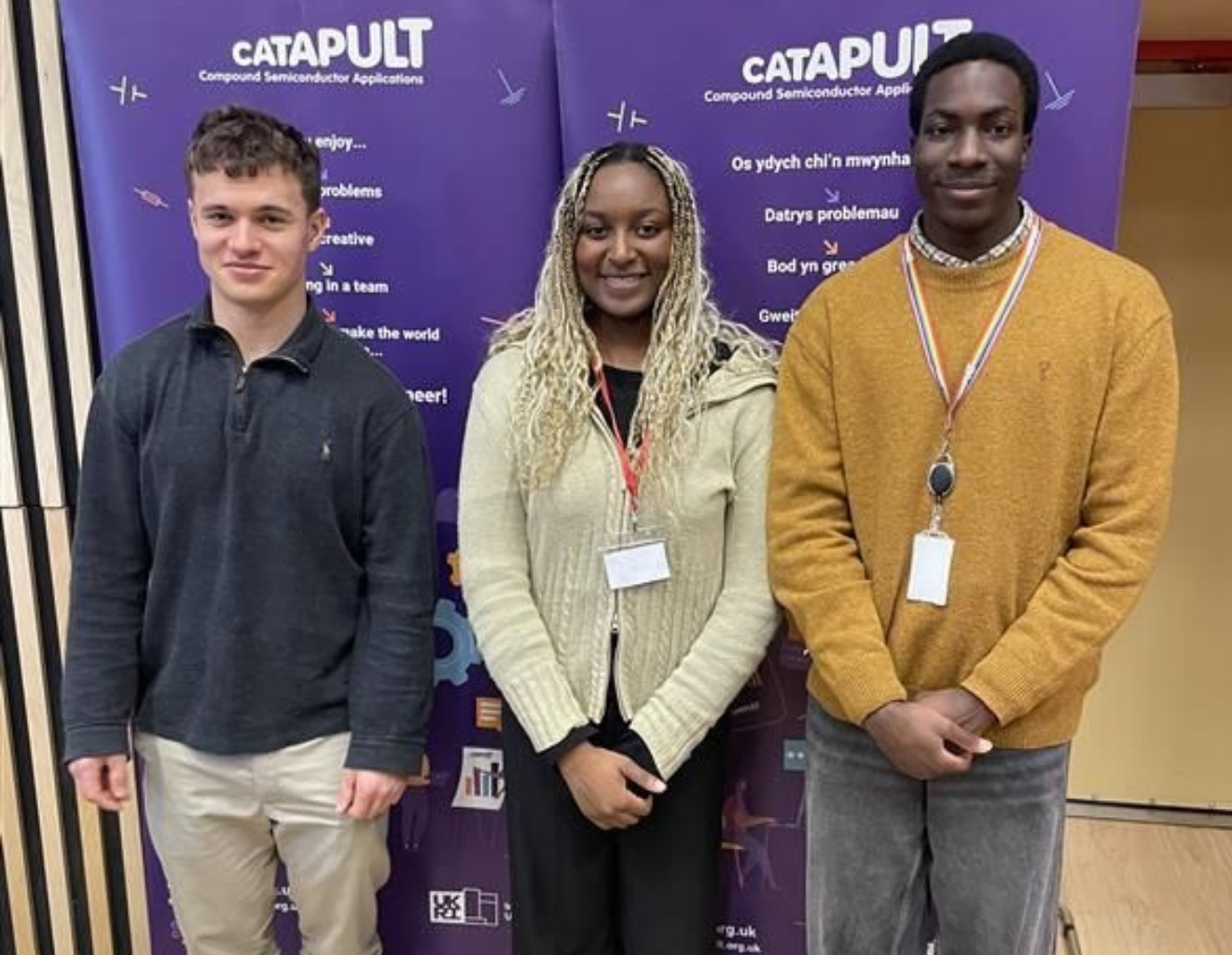 Three young adults stand smiling in front of Catapult banners at an indoor e-event. - CSA Catapult