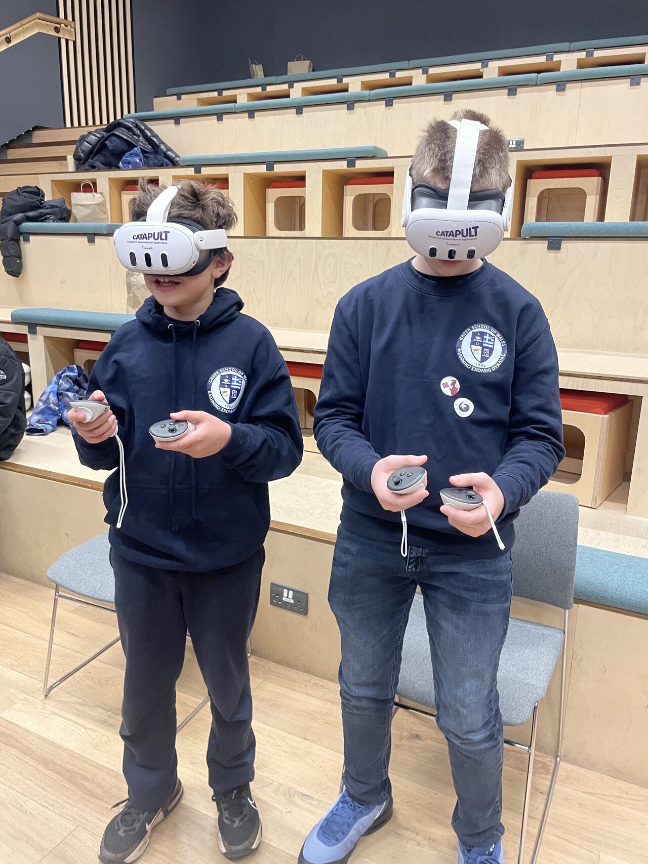 Two students wearing VR headsets and school uniforms stand indoors, each holding a VR controller and an e-device. - CSA Catapult