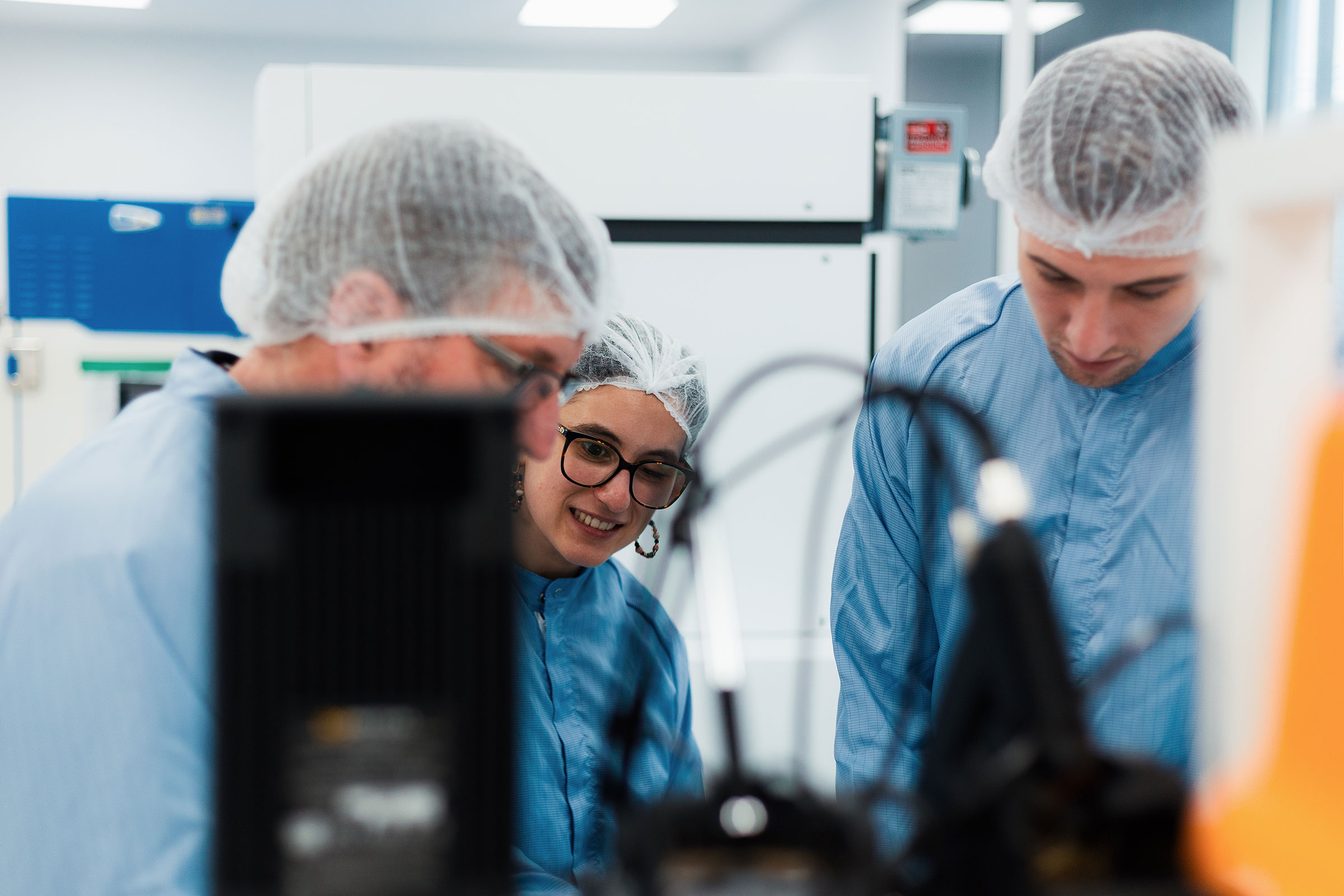 Three people in blue lab coats and hairnets work together in a laboratory setting with equipment visible. - CSA Catapult