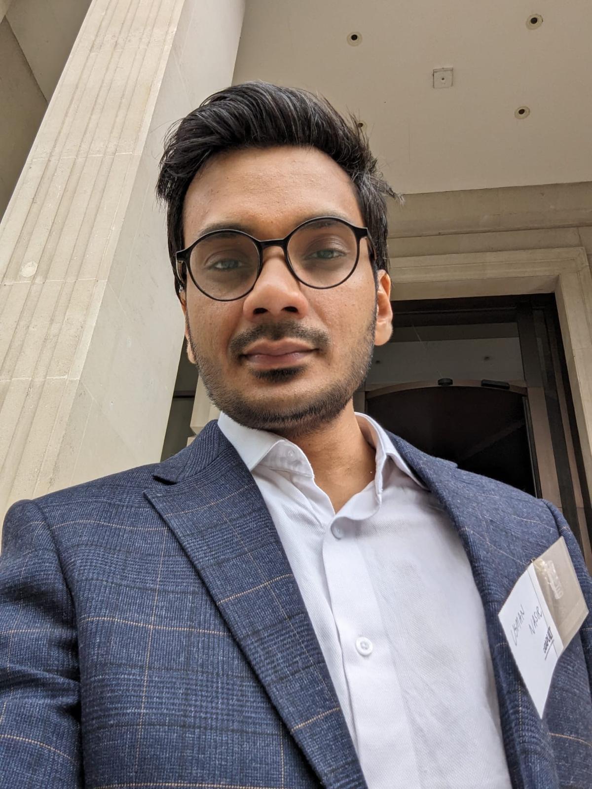A man in glasses and a suit stands outside a building, wearing an AI conference name tag and looking at the camera. - CSA Catapult