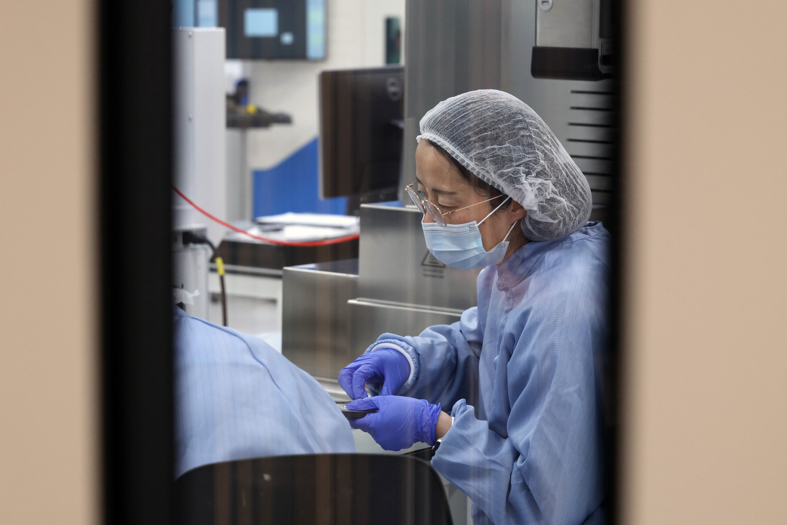 A lab technician in protective gear works with equipment in a laboratory, viewed through a window. - CSA Catapult