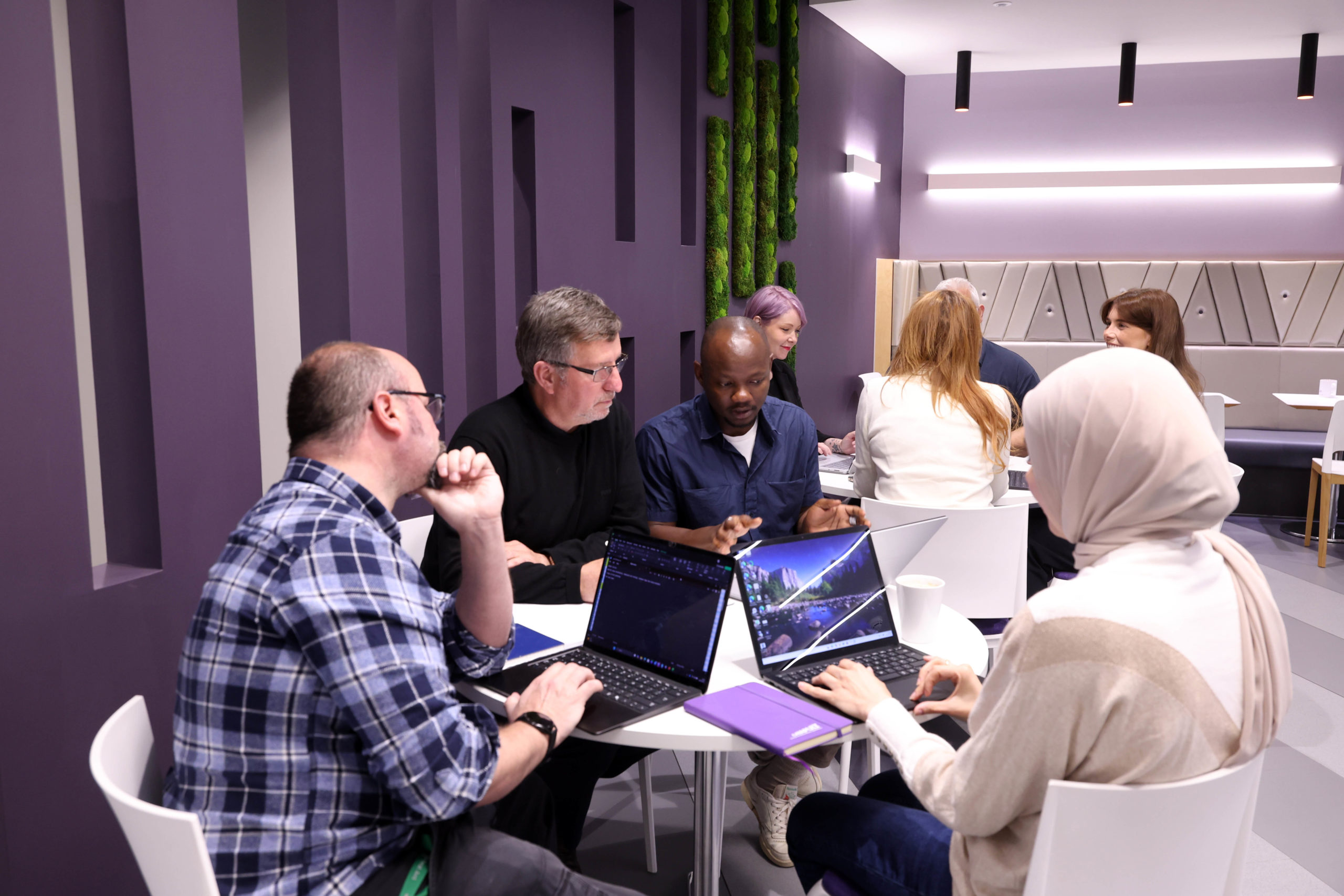 A group of people collaborate at a table with laptops in a modern, purple-themed office space. - CSA Catapult