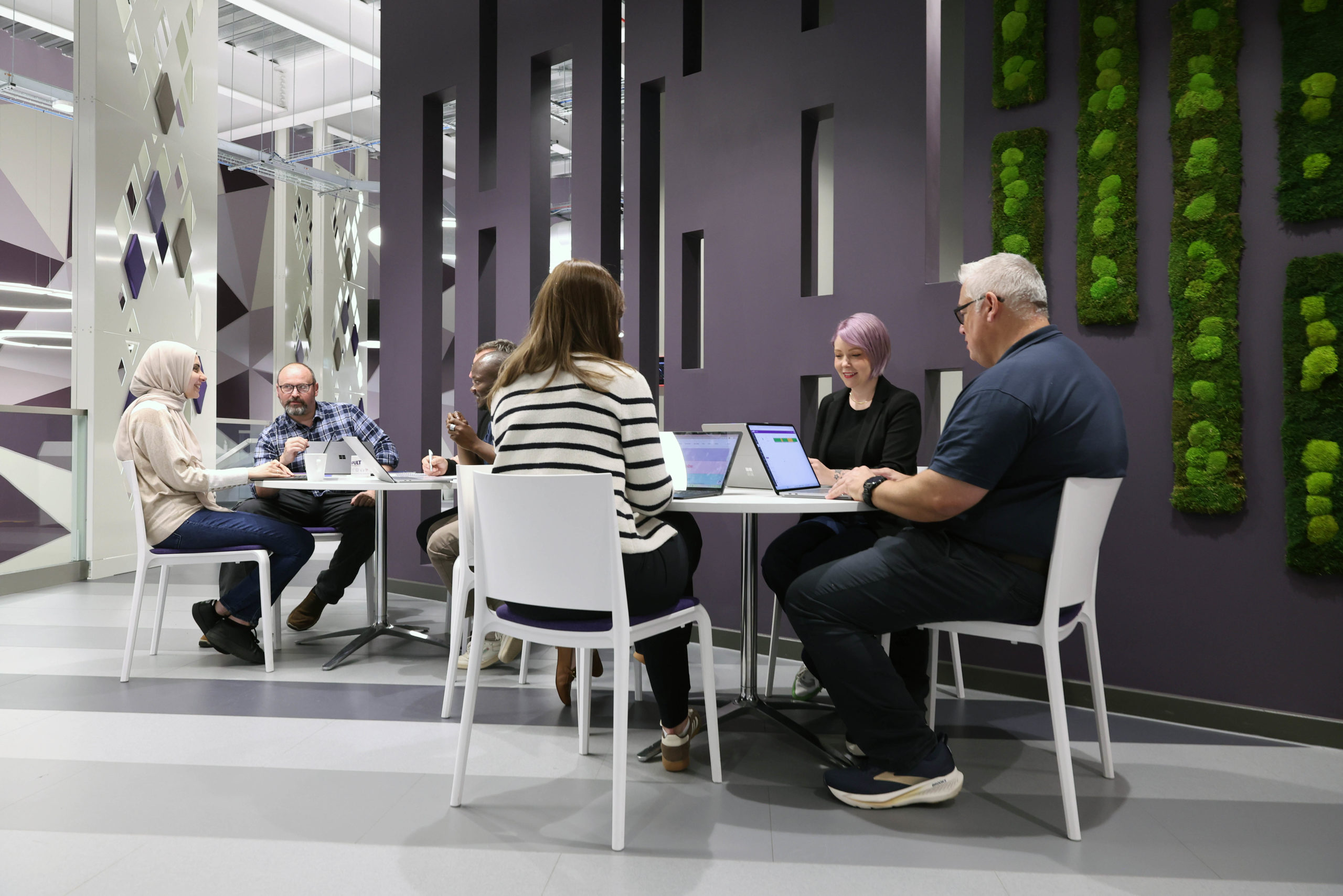 Six people sit at two round tables in a modern office, talking and working on laptops near green wall decor. - CSA Catapult