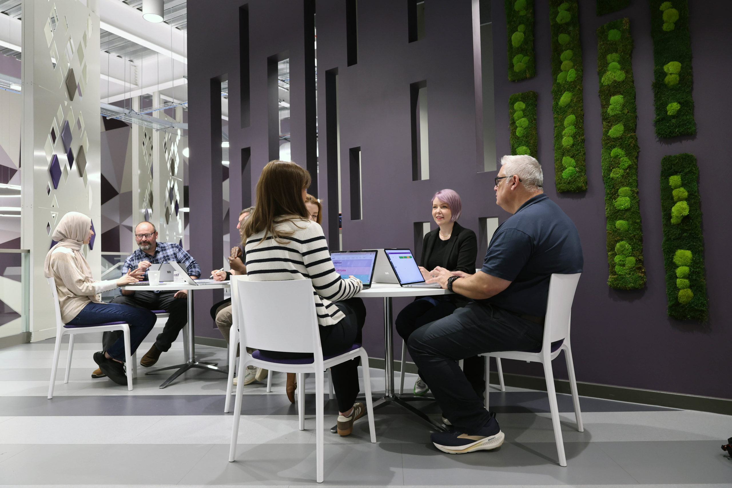 Six people sit around tables in a modern office, working and talking, with laptops and green wall decor behind them. - CSA Catapult