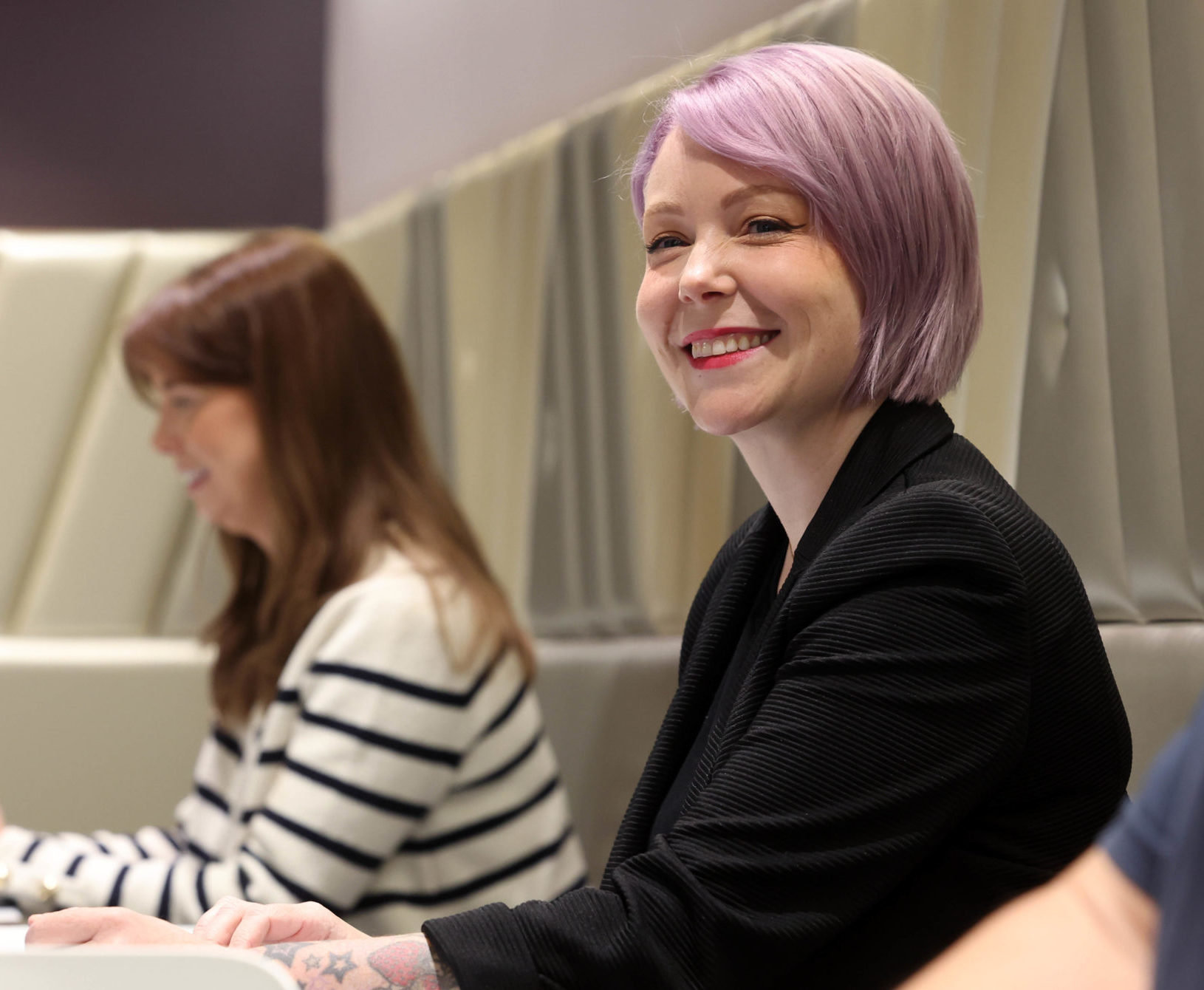 Smiling woman with purple hair works on a laptop, seated between two other people at a modern workplace. - CSA Catapult