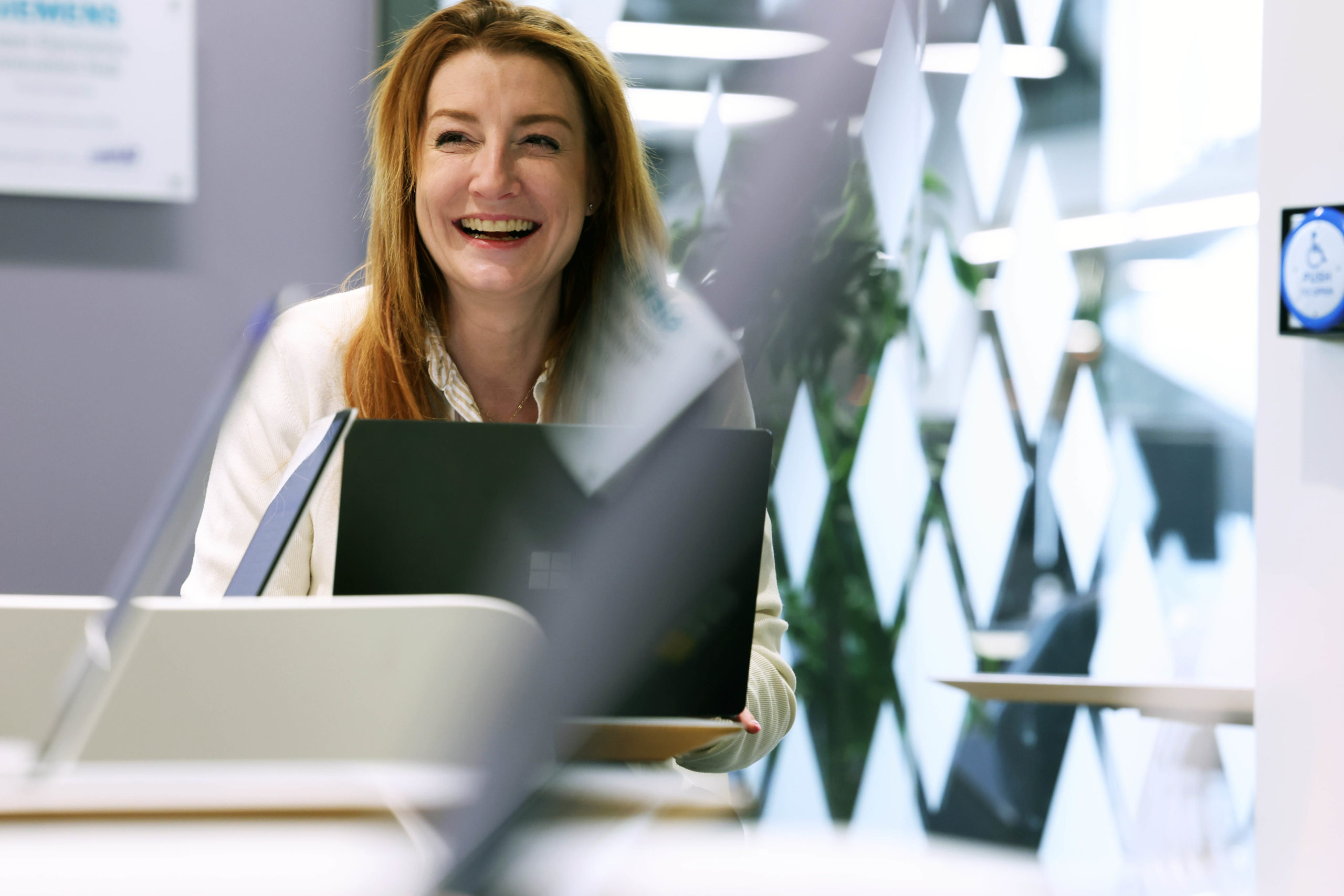 A woman smiling whilst working on a laptop in a bright, modern office space. - CSA Catapult