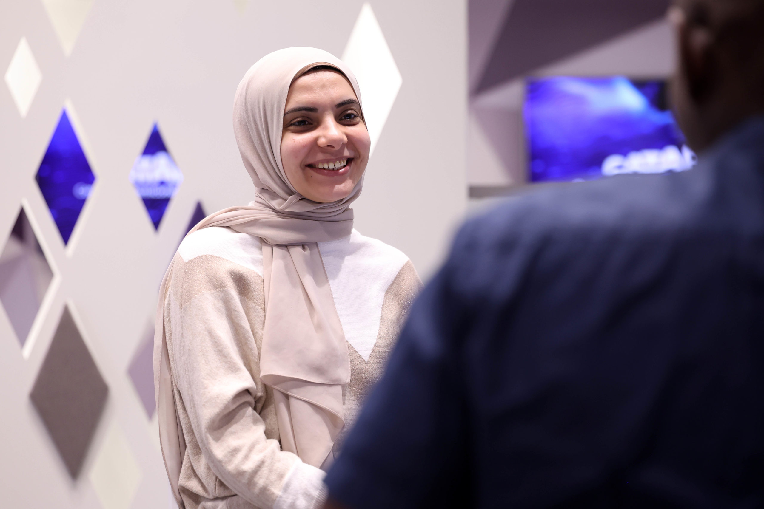 A smiling woman in a hijab talks to another person indoors with geometric décor in the background. - CSA Catapult