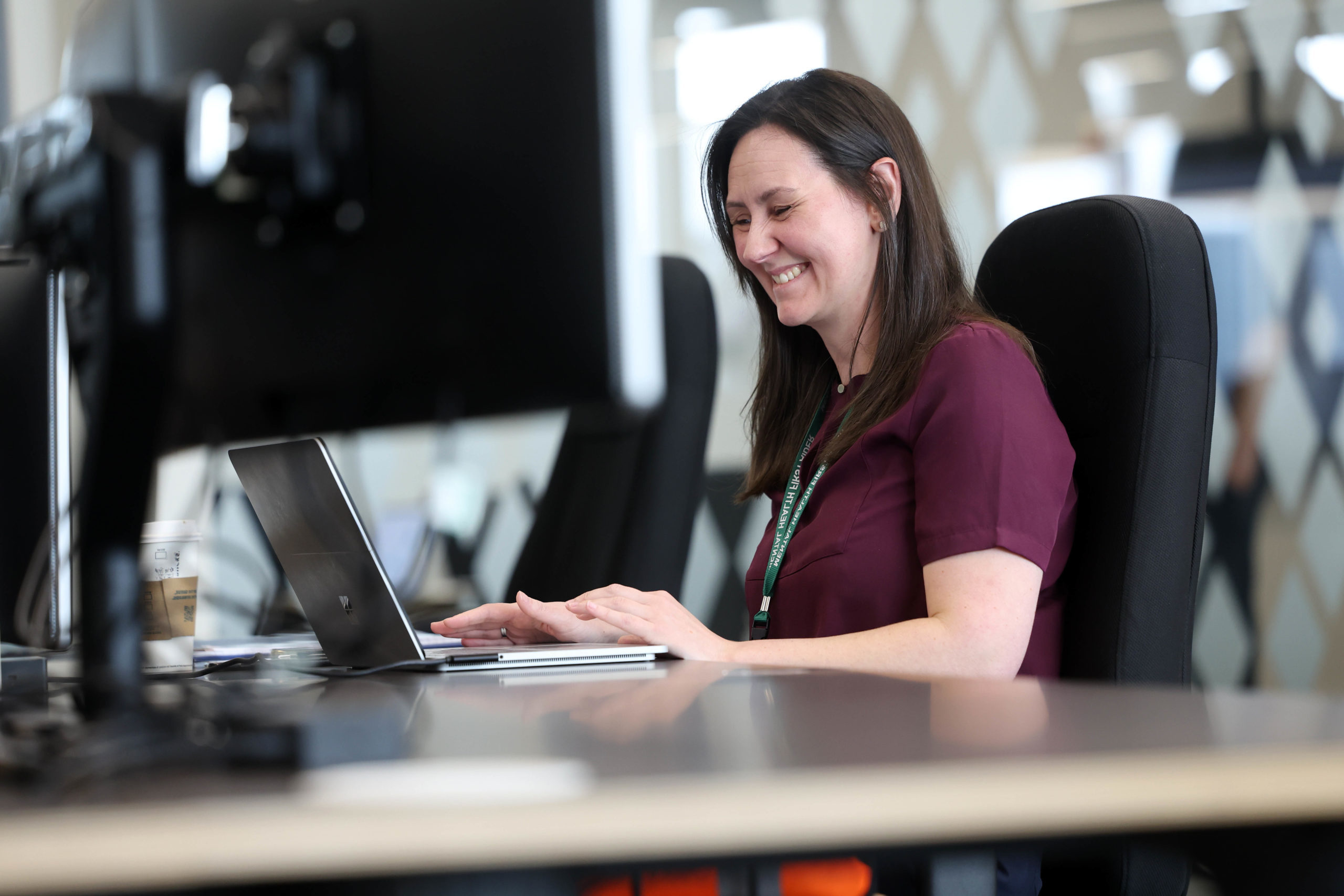 Woman smiling and working on a laptop at an office desk with computer monitors in the foreground. - CSA Catapult
