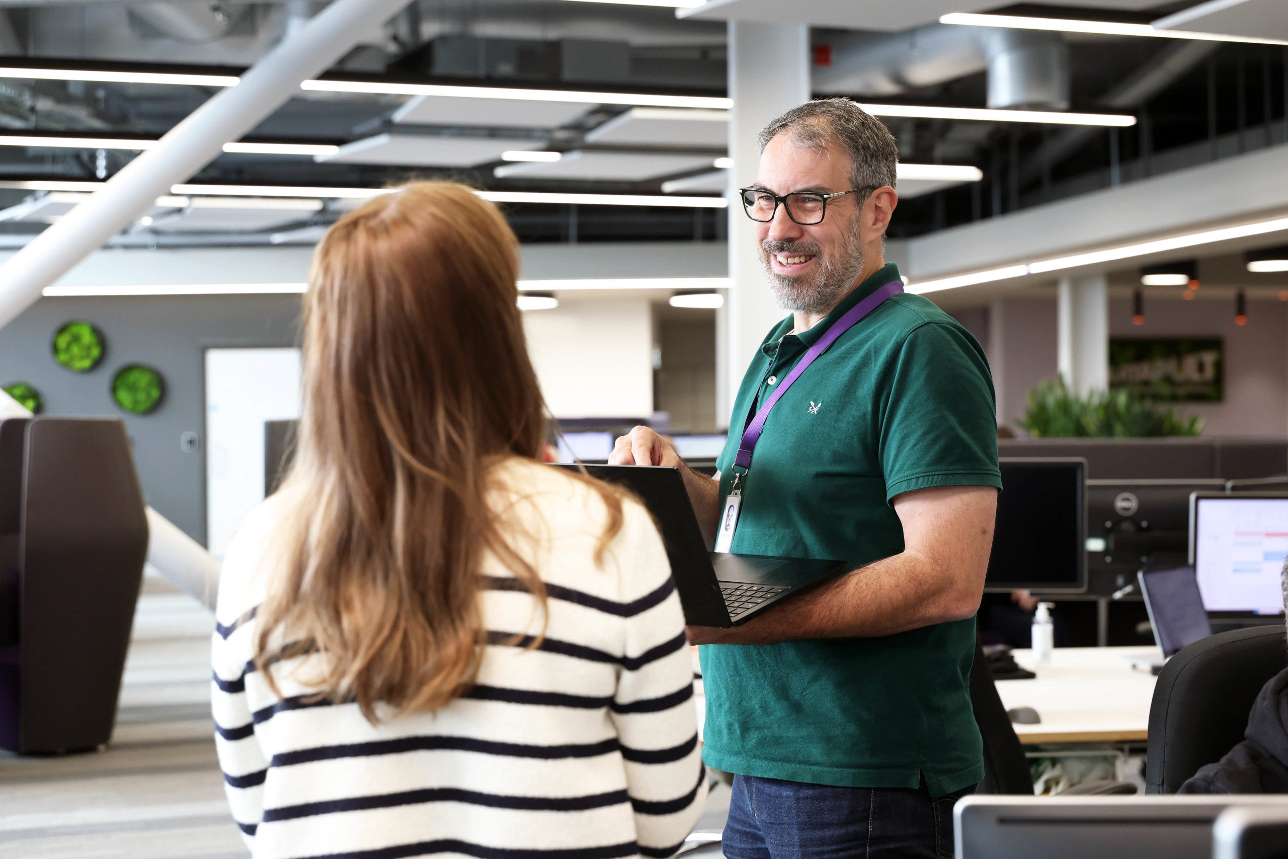 Man holding a laptop and smiling whilst talking to a woman in a modern office setting. - CSA Catapult