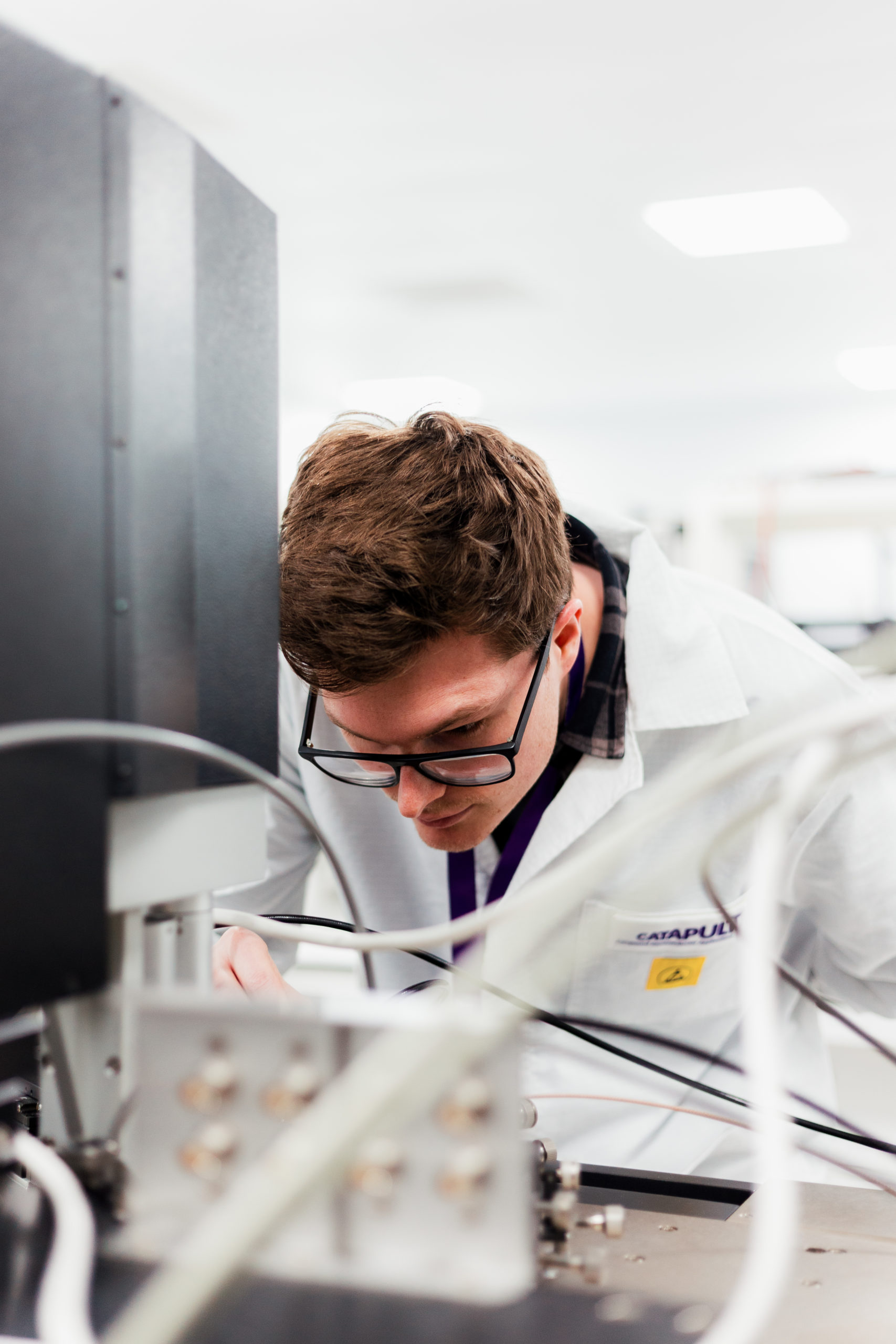 A person in a lab coat closely examines laboratory equipment with wires and machinery. - CSA Catapult