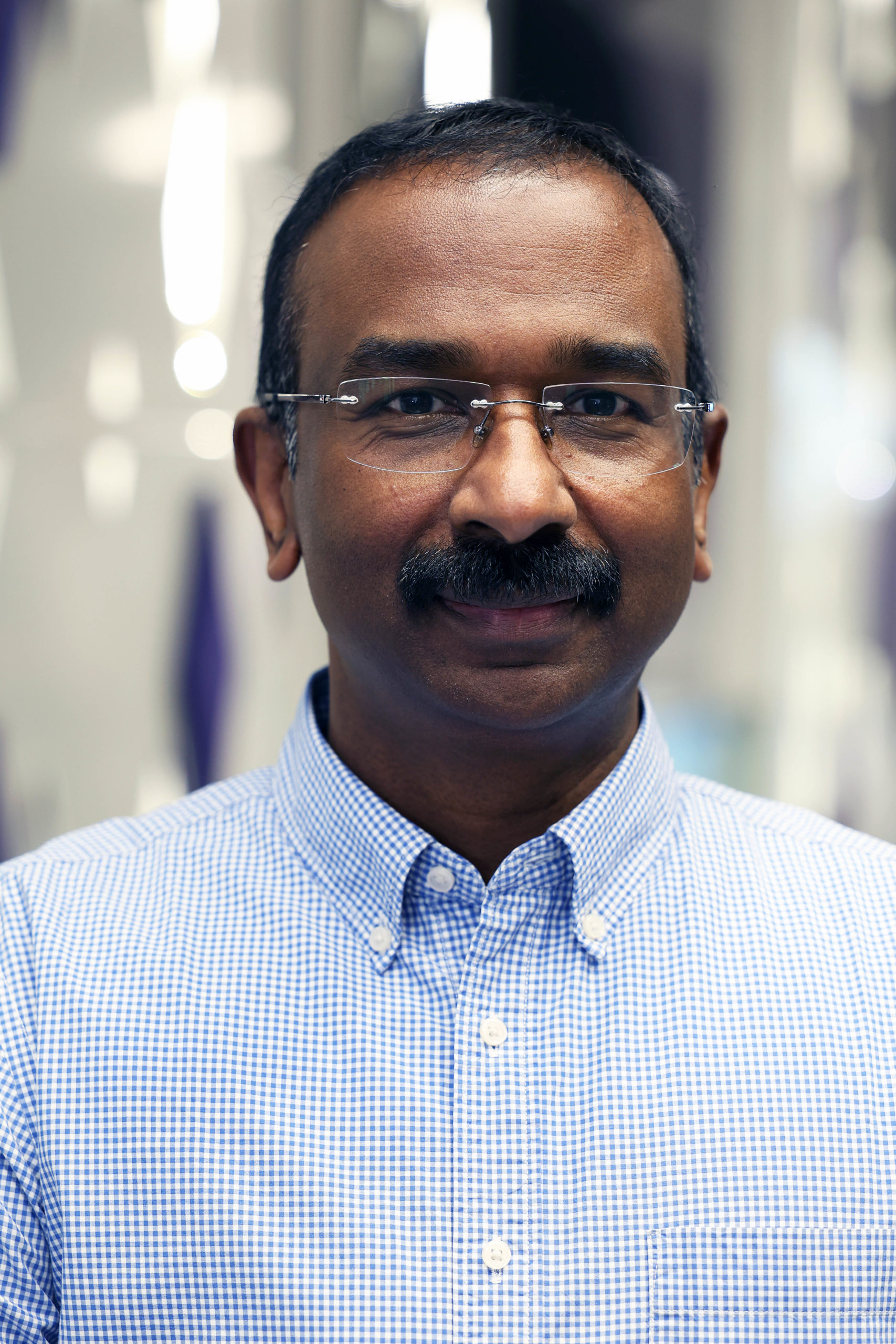 A man with glasses and a moustache smiles in a blue chequered shirt, standing indoors beside an AI device. - CSA Catapult