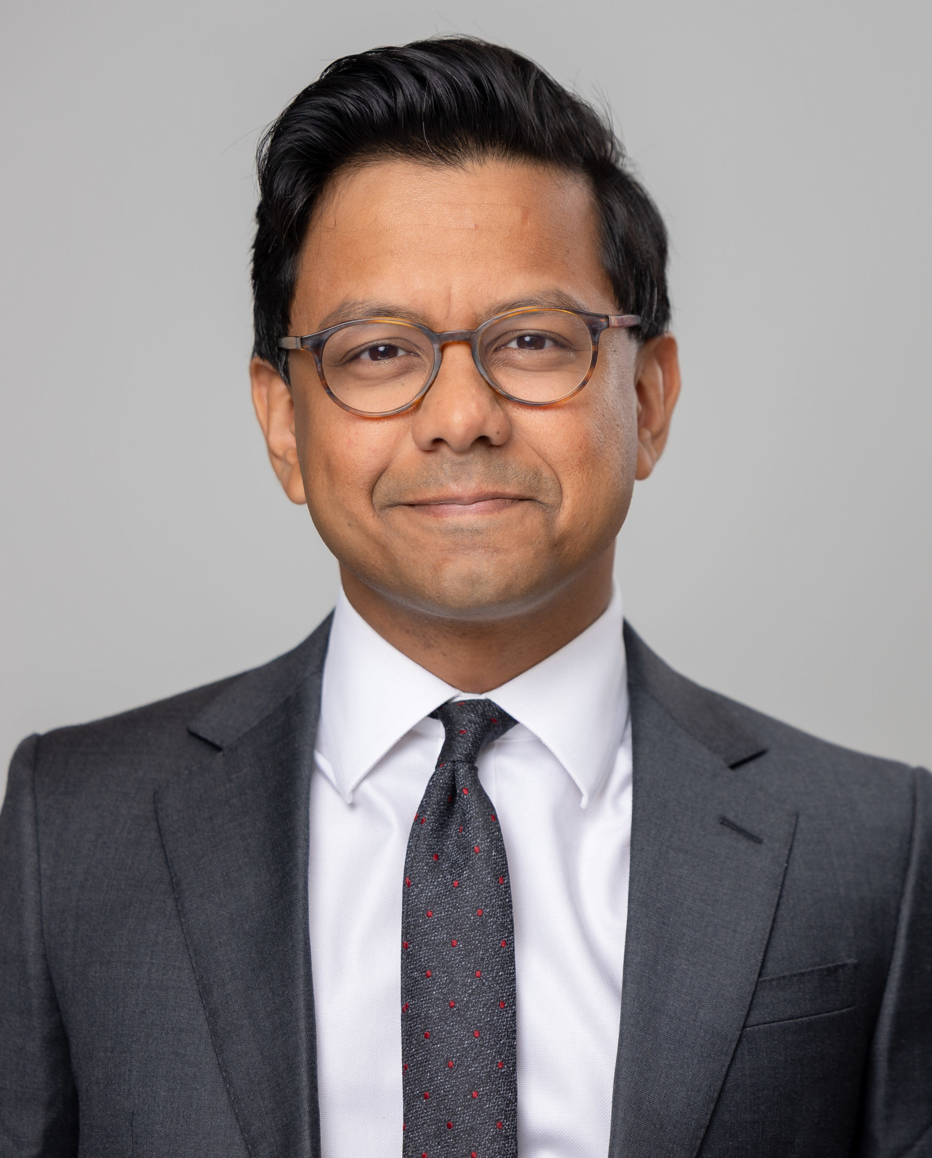 Man with short black hair and glasses, wearing a dark suit and tie, smiling slightly against a grey background. - CSA Catapult