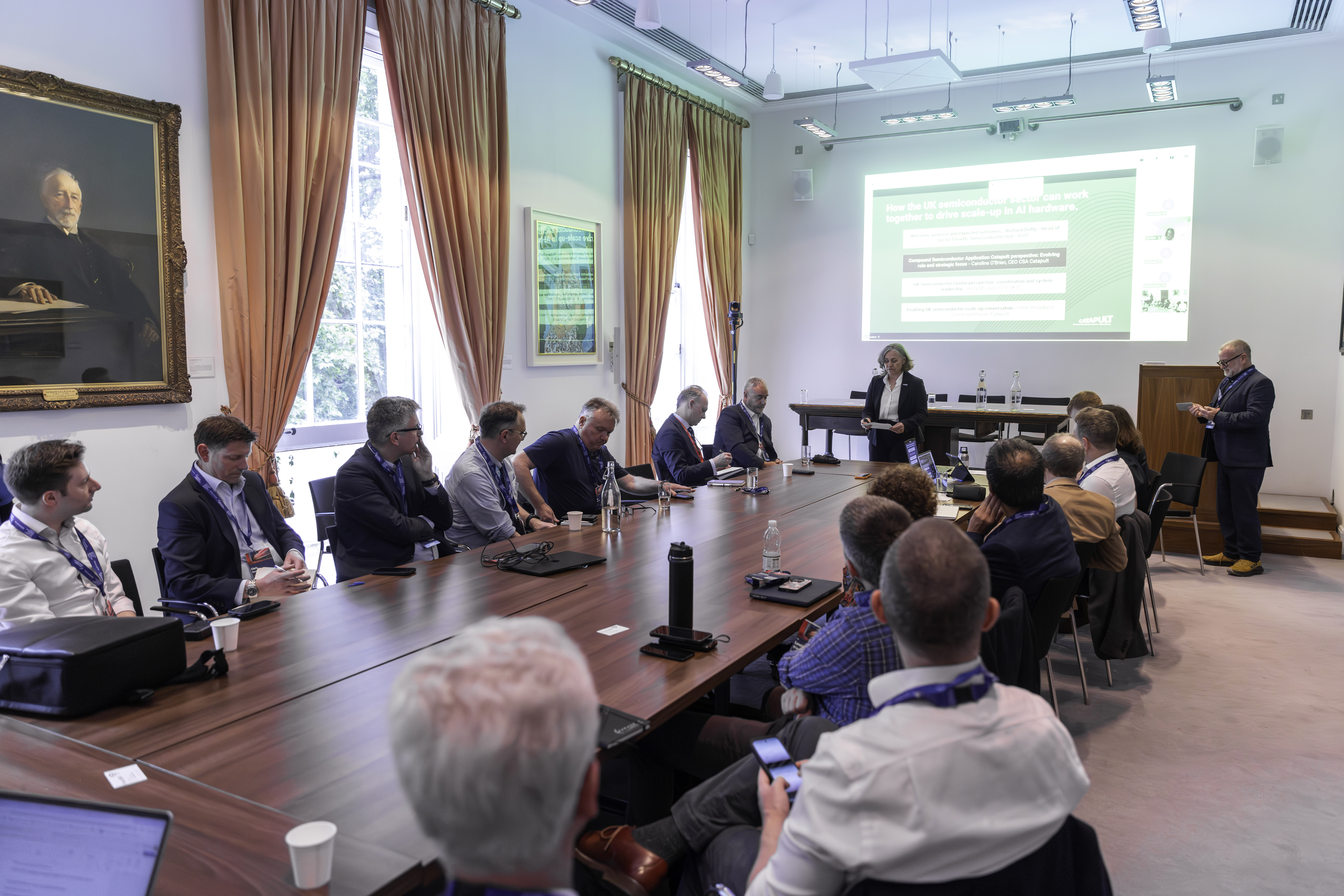 A group of people in business attire attend a presentation in a conference room with large windows and a screen. - CSA Catapult