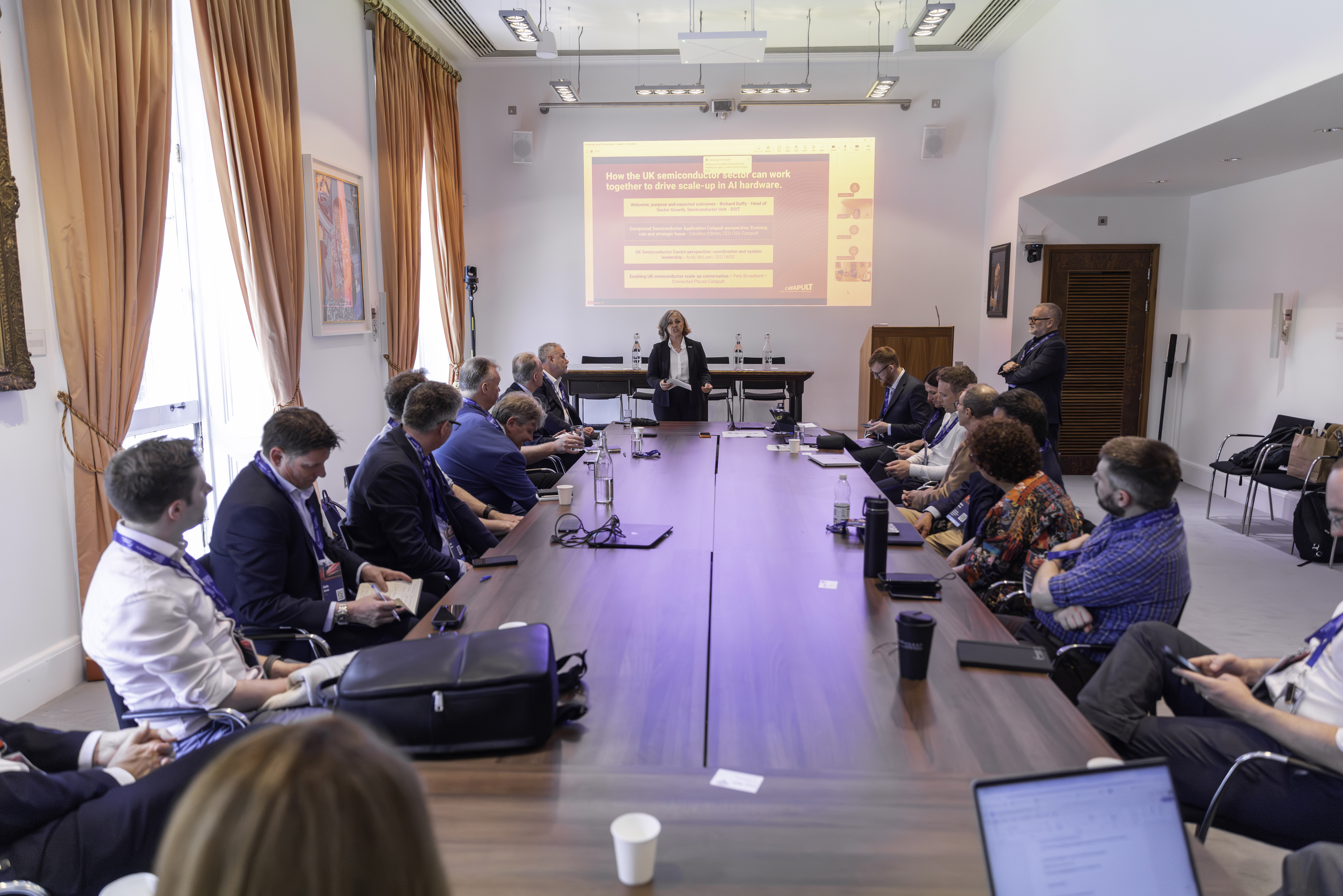 A woman presents to a group of people seated around a large conference table in a bright meeting room. - CSA Catapult
