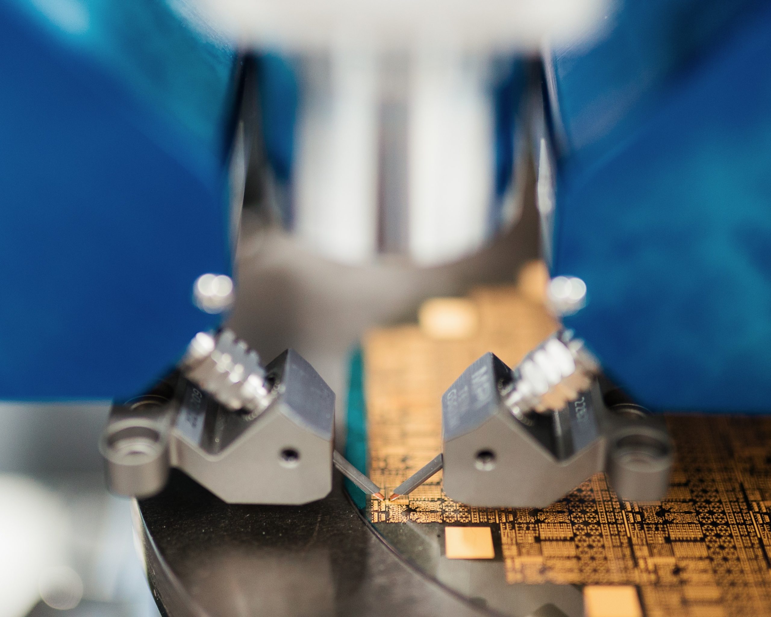 Close-up of a machine working on a brown circuit board, with two metal clamps and blue parts in view. - CSA Catapult